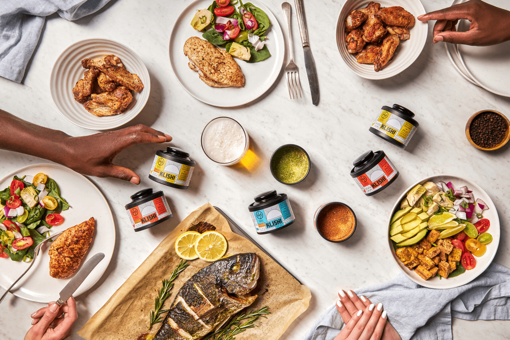 Top view of a kitchen counter with an arrangement of a diverse range of food and ingredients—chicken, fish, salads, and cutlery. In the center are multiple Smokey Hazel ‘KLISHI’ Jars.