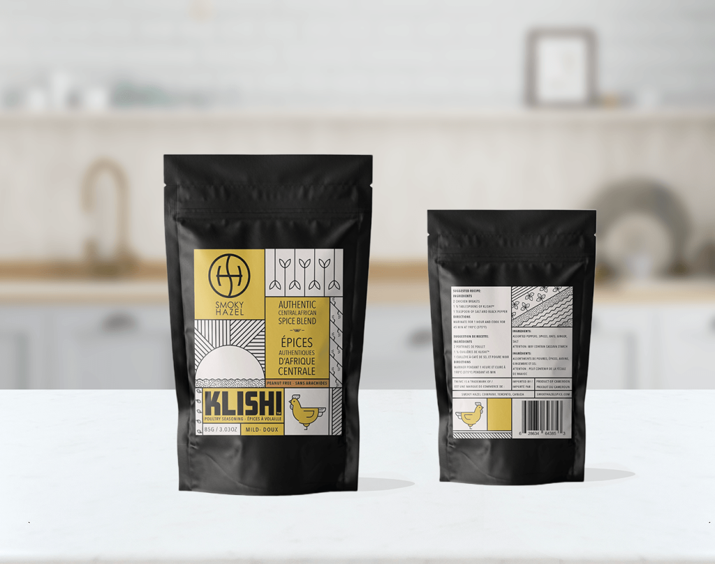 2 packets of Smokey Hazel ‘KLISHI’ spice blend sit on a countertop. One faces forward with graphics of a sun, a rooster, and text that reads "authentic blend" and the other backwards, showing nutritional information.