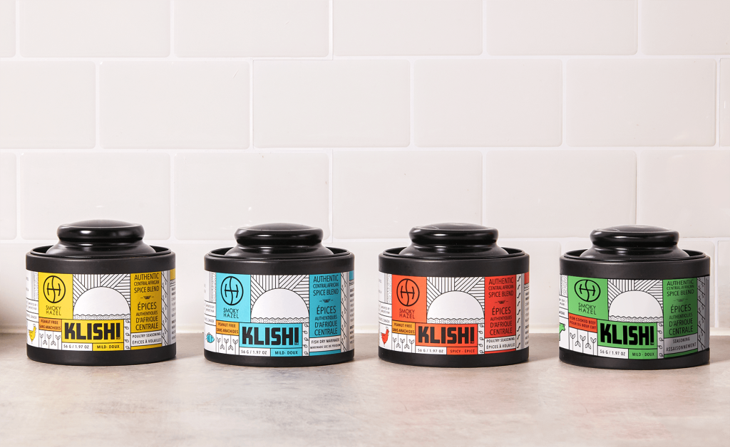 On a grey kitchen slab, 4 jars of the Smokey Hazel KLISHI spice blend in different flavors with different colored packaging – yellow, blue, red, and green. Each jar displays product branding, along with the Smokey Hazel logo.