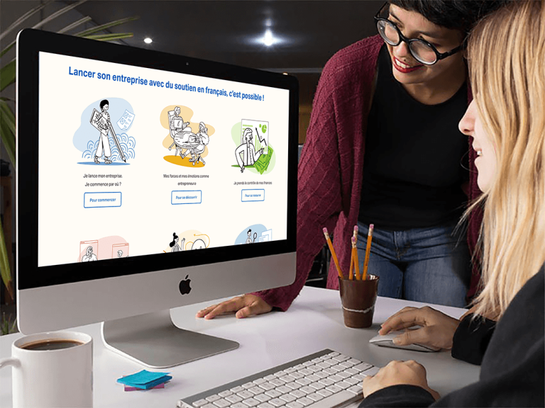 A woman sits at a computer desk and explores the Entrepreneurs 15:30 website while another woman stands over her, smiling at the screen.