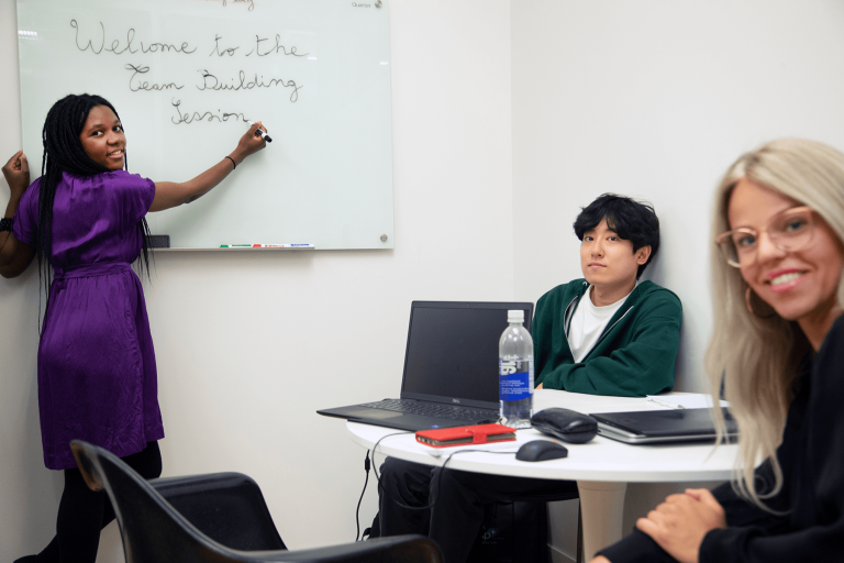 In a conference room with white-colored walls, Hermine Mbondo, founder and CEO of B4brand, in a purple dress and black stockings, writing “Welcome to the team building session” on a whiteboard while staring out to the camera. UX Designer, Daniel Lee, in a white shirt and green zipper hoodie, and a woman with platinum hair, sit around a round table and chairs with their laptops, looking out to the camera.