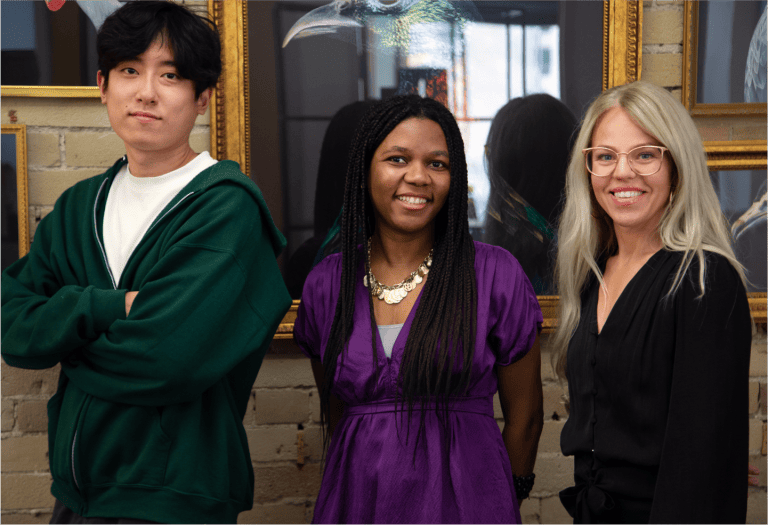 UX Designer Daniel Lee, Founder Hermine Mbondo, and a third person pose for a picture. They stand against a beige-colored brick wall with golden art frames in the background.