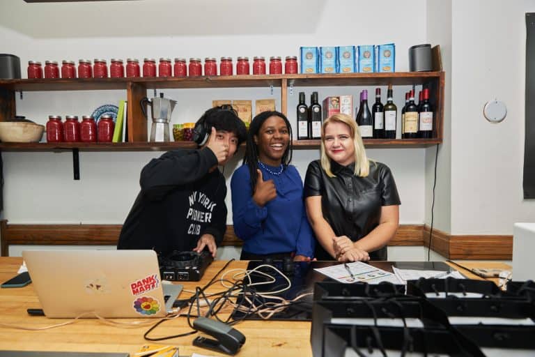UX Designer, Daniel Lee, in a black sweatshirt, wearing a headset and standing at a DJ booth, giving a thumbs-up. Next to him is B4brand Founder Hermine Mbondo in a blue shirt and black trousers, laughing and also showing a thumbs-up. A woman with blond hair and a nose ring stands by their side in a black leather dress.